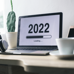 The image shows a computer screen with the words "2022 Loading..." and a loading bar on top of a work desk, in between a clock and cactus plant and a cell phone and cup of coffee.