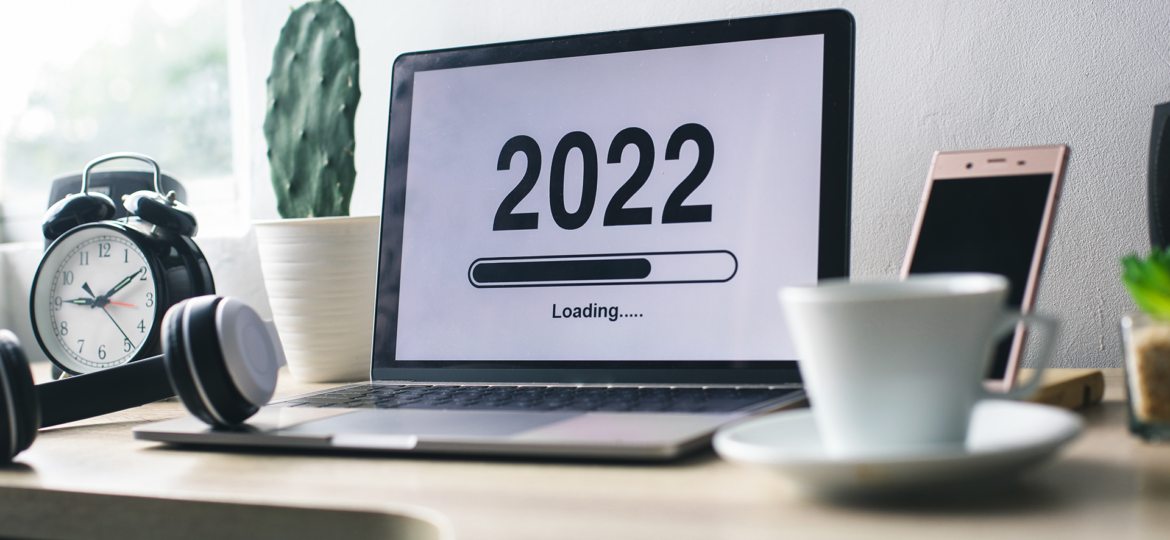 The image shows a computer screen with the words "2022 Loading..." and a loading bar on top of a work desk, in between a clock and cactus plant and a cell phone and cup of coffee.