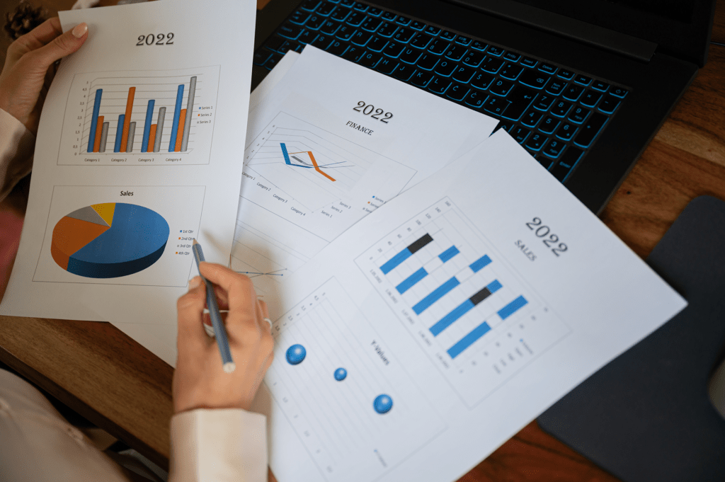 The image shows two hands, the right holding a pen and the left holding a sheet of paper with graphs that read "2022 Sales." A couple more sheets of paper with bar and line graphs read "2022 Finance" and are resting on top of a laptop. 